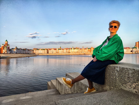 Young adult enjoys sunset overlooking waterfront cityscape with colorful buildings and calm riverの写真素材