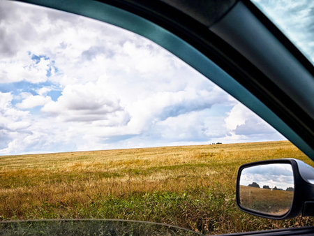 A serene view of rolling fields under a partly cloudy sky, framed by a car window on a peaceful day.の写真素材