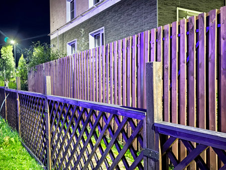 A wooden fence stands beside a well-kept garden, softly lit by streetlights in a tranquil neighborhood during nighttime.の写真素材