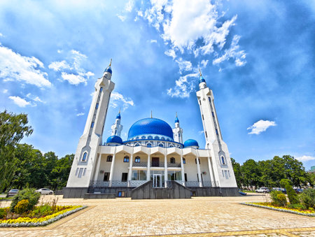 A grand mosque featuring a striking blue dome and tall, elegant minarets stands majestically against a bright blue sky, surrounded by greenery.の写真素材