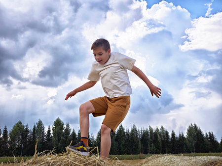 Boy jumping on haystack in a wide field under a dramatic sky during a sunny dayの写真素材