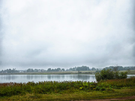 Fog rolls over a serene lake while grassy banks and distant houses create a peaceful scene on a cool morning.の写真素材