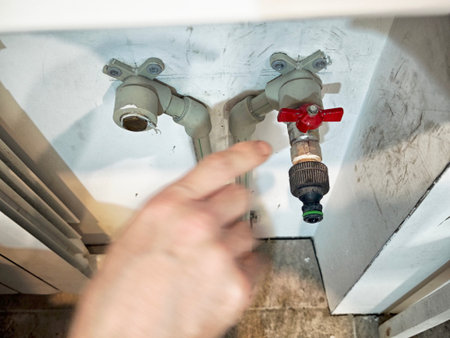 A hand is reaching to turn the red valve on a water pipe under a sink, indicating a plumbing adjustment or repair in a kitchen area.の写真素材