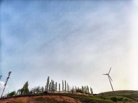 A wind turbine towers over a hilly area, surrounded by green trees, with a clear sky reflecting warm sunset hues.の写真素材