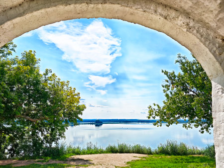 Calm river reflects a bright blue sky with clouds, seen through a stone arch surrounded by lush trees on a sunny afternoon.の写真素材