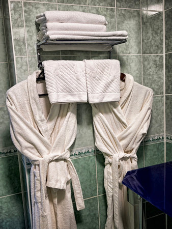 Two fluffy white bathrobes hang on hooks next to folded towels in a clean bathroom, creating a cozy...