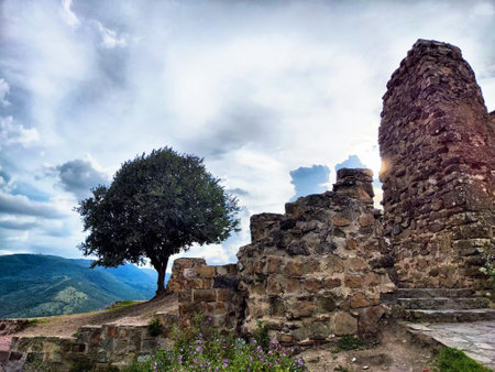 Ruins of an old structure stand on a hillside, with green mountains in the background and a single...