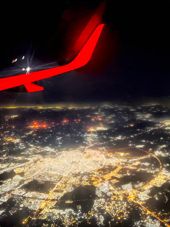 A colorful city skyline shines brightly at night from a plane, showcasing lights and urban life below, with a wing visible in the corner.の写真素材