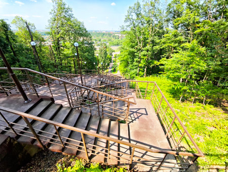 A serene staircase made of metal spirals down a hillside surrounded by lush green trees under clear...
