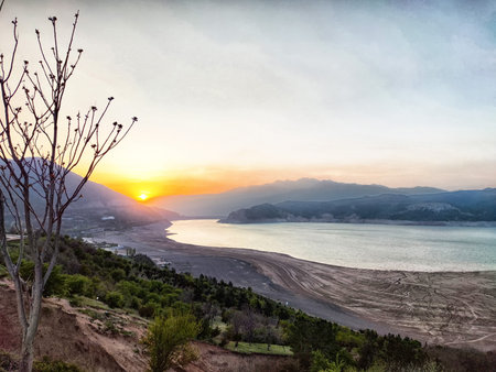 A beautiful sunset paints the sky orange as it sets over a peaceful lake surrounded by mountains and trees, creating a serene atmosphere.の写真素材