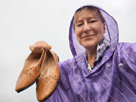 Blonde woman in a purple poncho smiles for a selfie, proudly holding stylish shoes in the rain on...