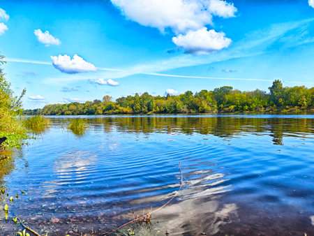 Calm river reflects blue sky and clouds while autumn trees line the shore, creating a serene nature...
