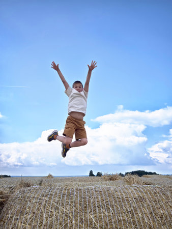 Child jumps joyfully in the air above a round hay bale under a bright blue sky during a sunny dayの写真素材
