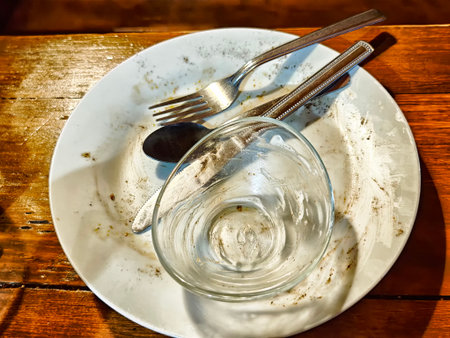Bright sunlight highlights a plate with leftover crumbs, a fork, knife, and an empty glass, indicating a finished meal.の写真素材
