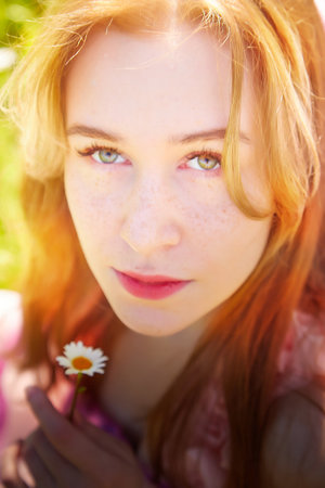 Young woman with orange hair holding a daisy in a sunny outdoor setting during springtimeの写真素材
