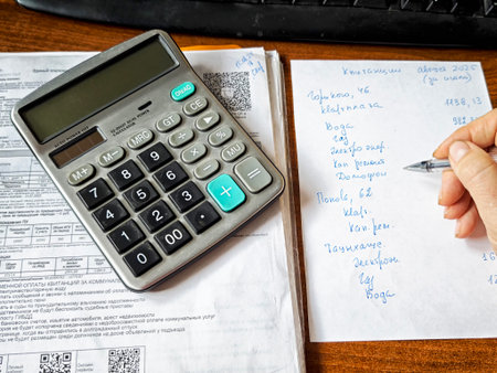 Kirov, Russia - August 15, 2025: A hand holds a pen to jot down figures while a calculator and sheets of paper are organized on a desk.のeditorial素材