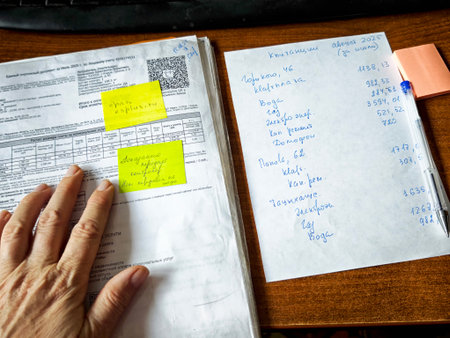 Kirov, Russia - August 15, 2025: A person is examining documents on a wooden desk filled with notes and calculations in a bright indoor setting.のeditorial素材