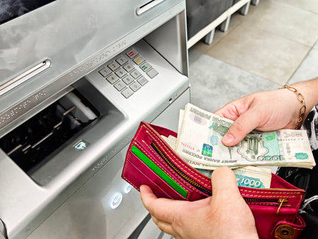 Maikop, Russia - June 04, 2025: A person is taking cash from an ATM while holding a wallet with Russian banknotes inside a contemporary bank.のeditorial素材