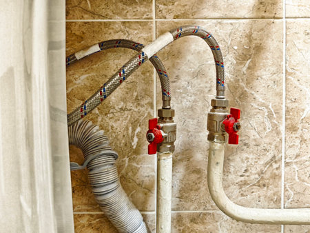 Close-up view of plumbing parts showing red valves and flexible pipes, highlighting a bathroom water supply setup.の写真素材