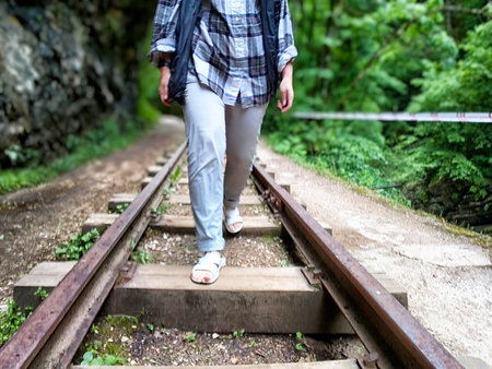 A person walks carefully on railway tracks surrounded by lush green trees and foliage under bright sunlight in a forest setting.の写真素材