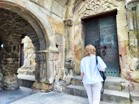 A woman stands before a large, ornate door within a weathered stone archway, observing the ancient...