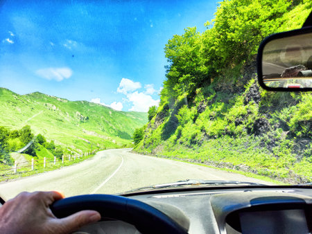 A person drives a car along a curved road surrounded by green hills and trees on a sunny day with...