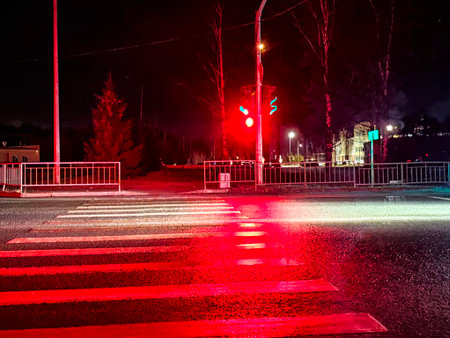 A red traffic light is visible above a crosswalk. The street is wet from rain, reflecting lights from the nearby buildings at night.の写真素材
