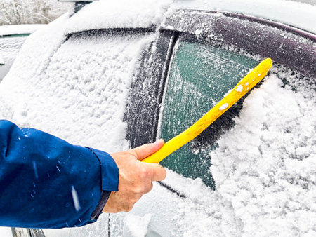 A man uses a snow brush to clear snow from a frozen vehicle windshield during a cold winter morning.