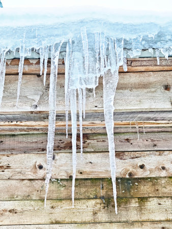 Long icicles form at the edge of a wooden roof, showing winter hazards and the risk of falling ice in cold weather conditions.の写真素材
