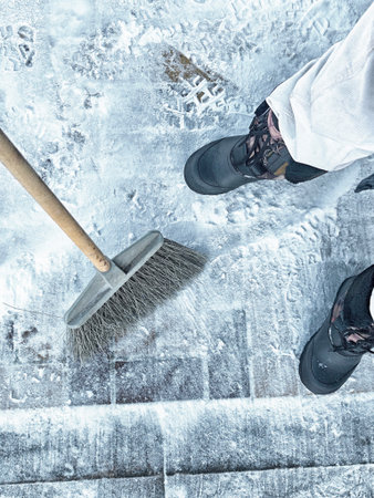 Individual sweeps snow off the paved walkway using a broom on a cold winter day while maintaining outdoor safety.の写真素材