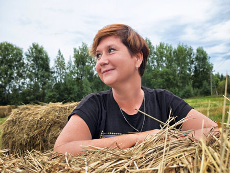 A mature woman smiles while relaxing on a hay bale in the countryside. She is enjoying her travel experience surrounded by nature.の写真素材
