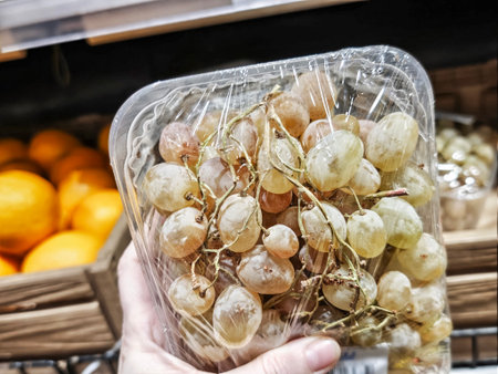 A hand holds a pack of grapes in a grocery store while selecting fresh food from the shelves near fruits.の写真素材