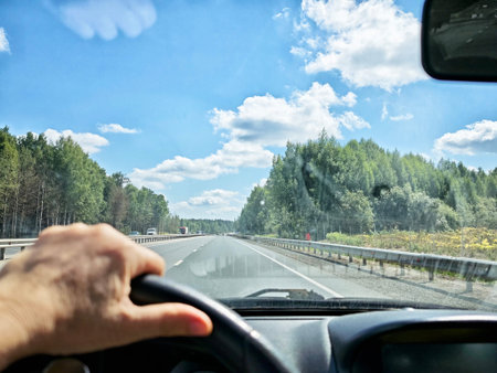 Hands grip the steering wheel as the car moves along an open highway, lined with lush trees under a...