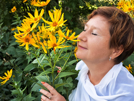 A middle-aged woman smiles while appreciating vibrant yellow flowers on her trip, embracing nature and adventure in a beautiful location.の写真素材