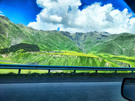 A rural road shows green hills and distant mountains under a bright sky with clouds. The landscape offers a glimpse into nature's beauty.の写真素材