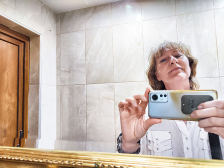 Woman stands in front of a mirror taking a selfie. She is capturing a moment of her daily life while...