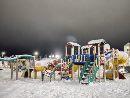 Children's playground covered in white snow during the night. Colorful play structures are visible...