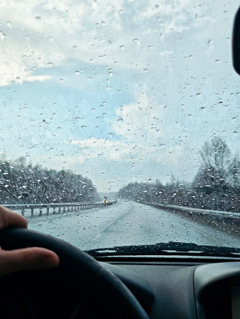 A road trip scene shows a driver in a car looking through a wet windshield at the road ahead. Rain falls with drops covering the glass.