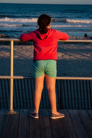 Young cute little girl on the boardwalk with back to camera looking towards the ocean surf