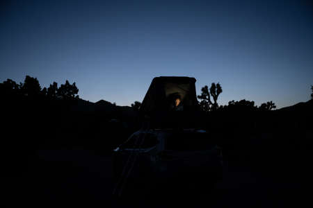Woman Sits In A Rooftop Tent and Works On A Computer At Sunset at campsite in Joshua Tree
