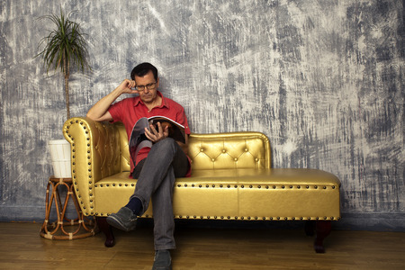mature man sits on golden leather sofa reading a magazineの写真素材