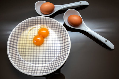 two egg yolks on a plate together with two shell eggs in spoons on black background