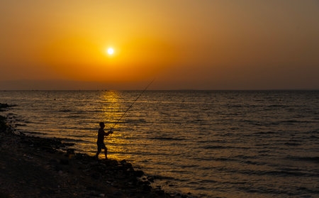 Silhouette of a man fishing on the beach at sunset.の写真素材