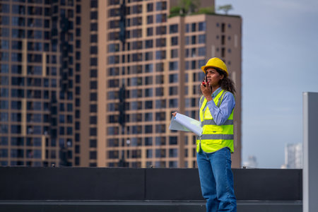 Front view of young African American female architect wearing safety vest and hardhat talking on...
