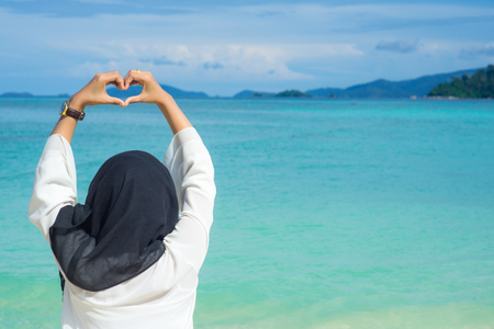 Beautiful young muslim woman black hijab lift hand showing heart shape at lipe thailand island with the turquoise ocean water.の写真素材