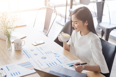 Asian beautiful young business woman in white shirt drinking latte art heart coffee working on desk with laptop and financial report document.have break time and resting after solving task.の写真素材