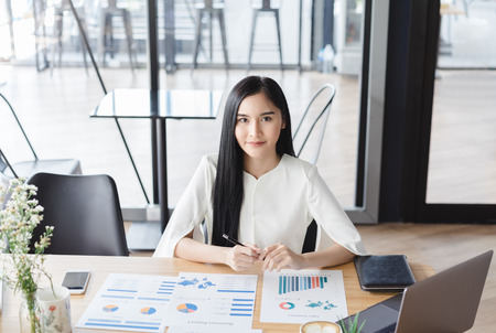 Asian beautiful young business woman in white shirt smiling working on desk with laptop and financial report document.の写真素材
