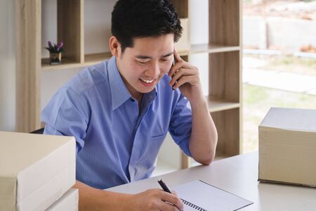 Young male startup small business owner receiving order from customer and talking by mobile phone. business owner working at home office. online shopping or freelance working concept.の写真素材