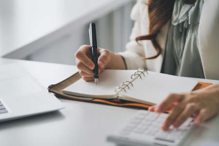 Cropped shot of woman hand writing on a notepad and using calculator in an modern office.の写真素材
