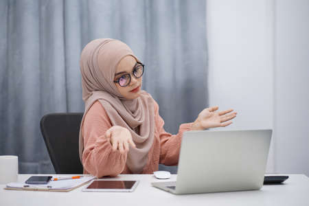 Attractive asian muslim woman in brown hijab having video call with friends or lover, using digital laptop at her home.の写真素材